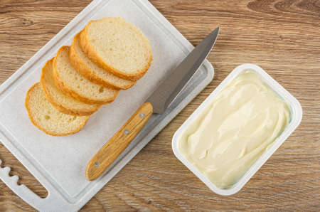 Slices of bread, kitchen knife on cutting board, plastic box with cream cheese on wooden table. Top...