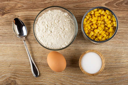 Ingredients for corn pancakes. Bowl with flour, jar of sweet corn, brown egg, salt cellar on wooden...