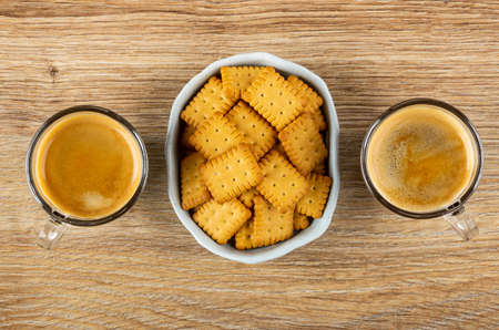 Two transparent cups with coffee espresso and light-blue bowl with small salted crackers on wooden table. Top viewの写真素材