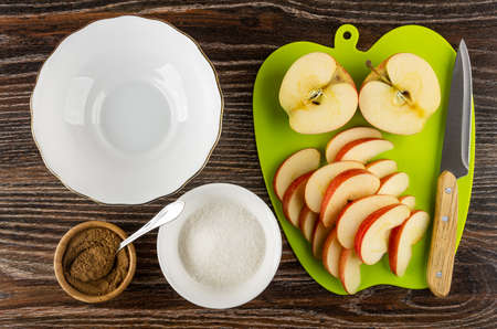 Empty glass bowl, spoon in bamboo bowl with ground cinnamon, bowl with sugar, pieces of apples on green cutting board on wooden table. Top viewの写真素材