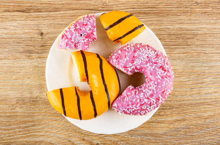 Sections of pink glazed donut with sprinkles and yellow donut with chocolate stripes in white plate on wooden table. Top viewの写真素材