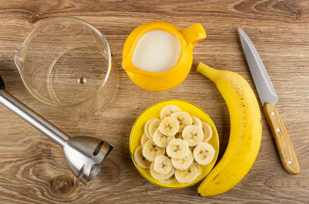 Empty transparent glass, electric blender, yellow pitcher with milk, slices of peeled banana in saucer, banana, kitchen knife on wooden table. Top viewの写真素材