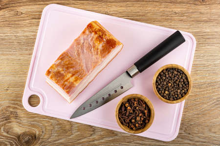 Piece of smoked brisket, kitchen knife, bamboo bowls with black peppercorn and clove spice on pink...