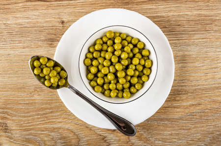 White glass bowl with canned green peas, metallic spoon with green peas in plate on brown wooden...