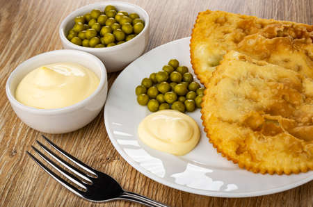 Bowls with green peas and mayonnaise, white plate with two fried cheburek, green peas and...