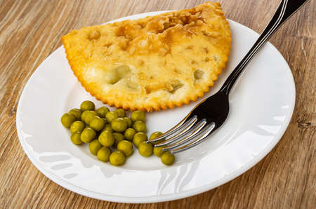 White plate with one fried cheburek, green peas and fork on brown wooden table