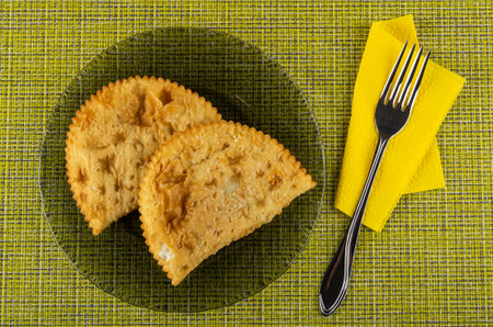 Two fried cheburek in transparent gray plate, fork on paper napkin on yellow mat. Top view