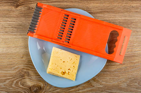Piece of cheese, plastic orange grater in light-blue plate on brown wooden table. Top view