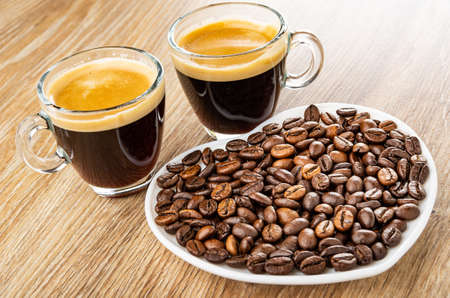 Two transparent cups with black coffee, roasted coffee beans in white saucer in heart shape on brown...