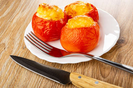 Baked tomatoes with filling from egg and cheese in white plate, kitchen knife, fork on brown wooden tableの写真素材