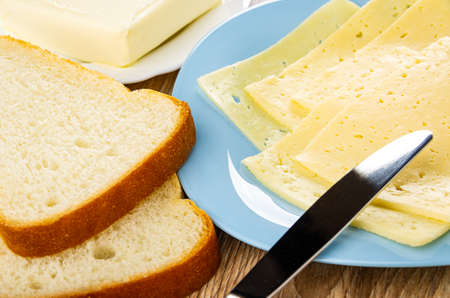 Piece of butter in white plate, slices of wheat bread, table knife on light-blue glass plate with thin slices of cheese on wooden tableの写真素材