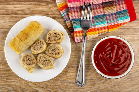 Whole roll pancake, pieces of roll pancake with meat filling in white plate, fork on checkered napkin, bowl with tomato ketchup on wooden table. Top viewの写真素材