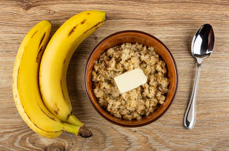 Two bananas, dairy oatmeal with piece of butter in brown bowl, spoon on wooden table. Top viewの写真素材