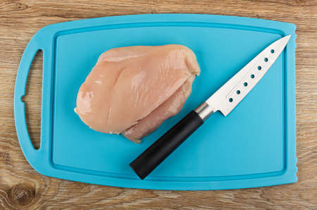 Piece of raw chicken fillet, kitchen knife on blue plastic cutting board on wooden table. Top viewの写真素材