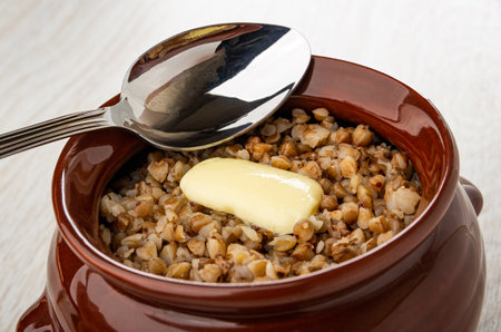 Metallic spoon, cooked buckwheat porridge with butter in brown ceramic potの写真素材