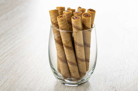 Brown striped wafer rolls with chocolate filling in transparent drinking glass on wooden tableの写真素材