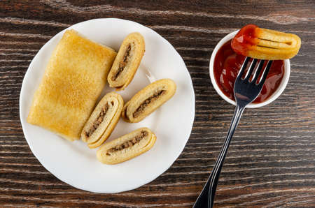 Whole pancake roll, pieces of fried pancake with meat filling in white plate, piece piece of pancake on fork above bowl with ketchup on dark wooden table. Top viewの写真素材