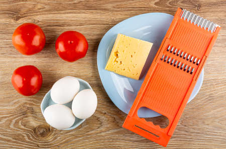 Few red tomatoes, white eggs, piece of cheese, plastic orange grater in light-blue plate on brown wooden table. Top viewの写真素材