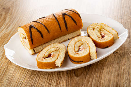 Piece of brown striped swiss roll, slices of roll with cream and jam in white dish on wooden table