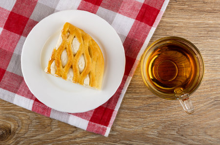 Piece of wicker sweet pie with cottage cheese in white plate on checkered napkin, transparent cup with hot tea on wooden table. Top viewの写真素材
