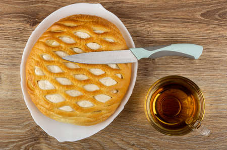 Kitchen knife on wicker sweet pie with cottage cheese in white dish, transparent cup with hot tea on...