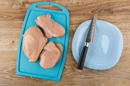Three pieces of raw chicken fillet on blue plastic cutting board, kitchen knife in glass plate on wooden table. Top viewの写真素材
