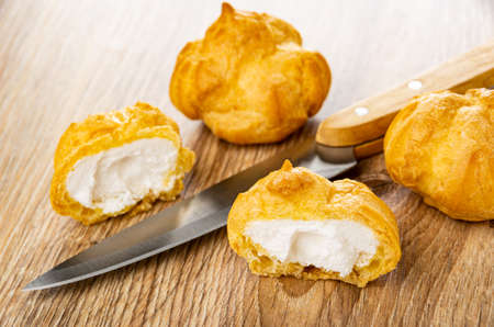 Whole profiteroles, halves of profiteroles with whipped cream, kitchen knife on wooden table