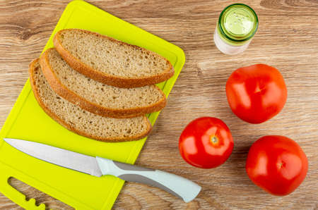 Slices of rye bread, kitchen knife on green cutting board, salt shaker, ripe red tomatoes on wooden...