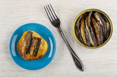 Sandwich with sprats in blue saucer, fork, metallic jar with sprats on wooden table. Top viewの写真素材