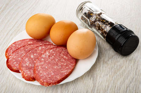 Sliced smoked-boiled sausage, brown eggs in white plate, transparent jar with condiment on wooden tableの写真素材