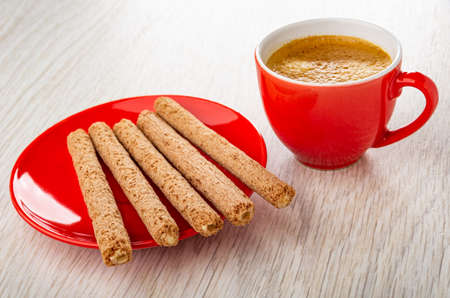 Crusty sticks with cream filling in red saucer, coffee espresso in cup on wooden table