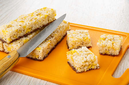Few cereal bars with coconut and chocolate, kitchen knife, pieces of cereal bar on orange cutting...
