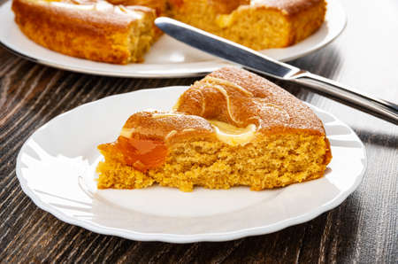Cake with cream and jam in white glass dish, table knife, slices of cake in plate on dark wooden...