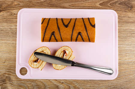 Brown striped swiss roll, table knife on slices of roll on pink cutting board on wooden table. Top viewの写真素材