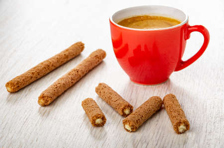 Brown sticks with cream filling, red cup with coffee espresso, broken crusty sticks on wooden table