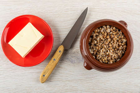 Piece of butter in red saucer, kitchen knife, brown ceramic pot with cooked buckwheat porridge on...