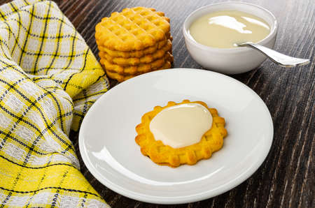 Checkered napkin, stack of waffle cookies, teaspoon in bowl with condensed milk,cookie poured condensed milk in white saucer on dark wooden tableの写真素材