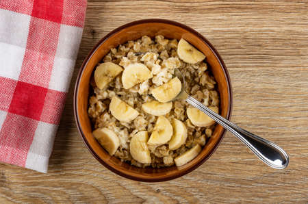 Checkered napkin, spoon in bowl with dairy oatmeal and slices of banana on wooden table. Top viewの写真素材