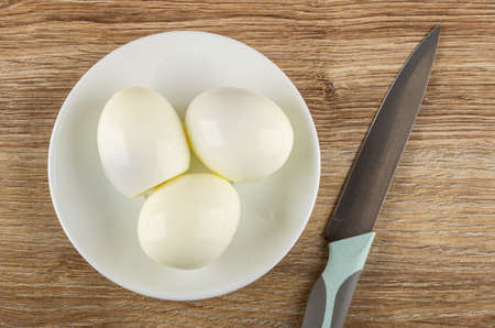 Three boiled peeled eggs in white saucer, kitchen knife on wooden table. Top viewの写真素材