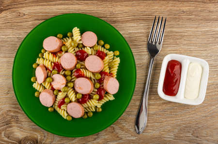 Green glass plate with boiled pasta fusilli, fried sausages and tomato sauce, fork, partitioned sauce boat with ketchup and mayonnaise on wooden table. Top viewの写真素材
