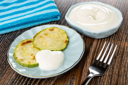 Striped napkin, bowl with sour cream, slices of fried vegetable marrow with sour cream in blue saucer, fork on dark wooden tableの写真素材