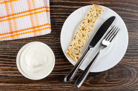 Checkered napkin, glass bowl with sour cream, pancake roll with filling, table knife and fork in...