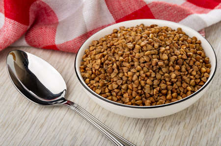 Checkered napkin, metallic spoon, dry buckwheat in white glass bowl on wooden table