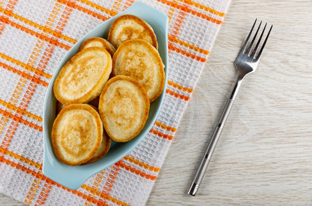 Small homemade pancakes in blue oval plate on checkered napkin, fork on wooden table. Top viewの写真素材