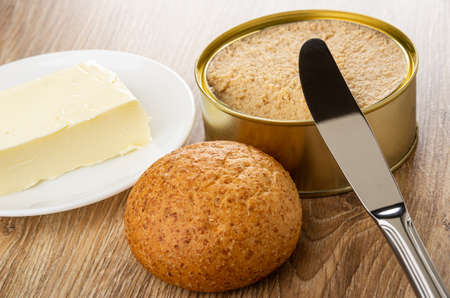 Piece of butter in white saucer, table knife on opened metallic jar with canned sterilized cod caviar, small cereal bun on wooden tableの写真素材