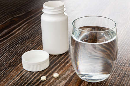 Opened plastic jar, lid, two white small round tablets, glass of water on dark wooden tableの写真素材