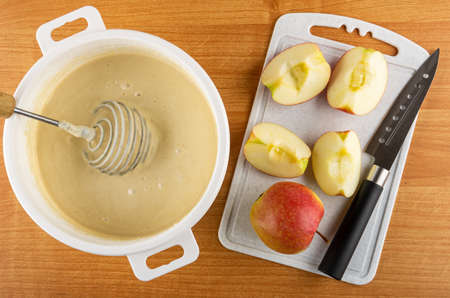 Whisk in white plastic pan with dough, pieces of red apples, kitchen knife on cutting board on wooden table. Top viewの写真素材