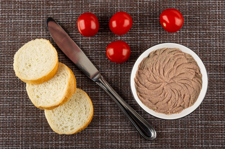 Slices of wheat bread, table knife, few tomatoes cherry, white bowl with liver pate on dark mat. Top...