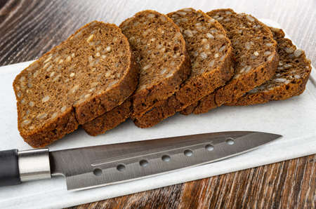 Slices of rye bread with sunflower seeds, kitchen knife on plastic cutting board on dark wooden...