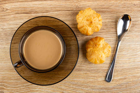 Brown transparent cup of coffee with milk on saucer, profiteroles with whipped cream, teaspoon on...